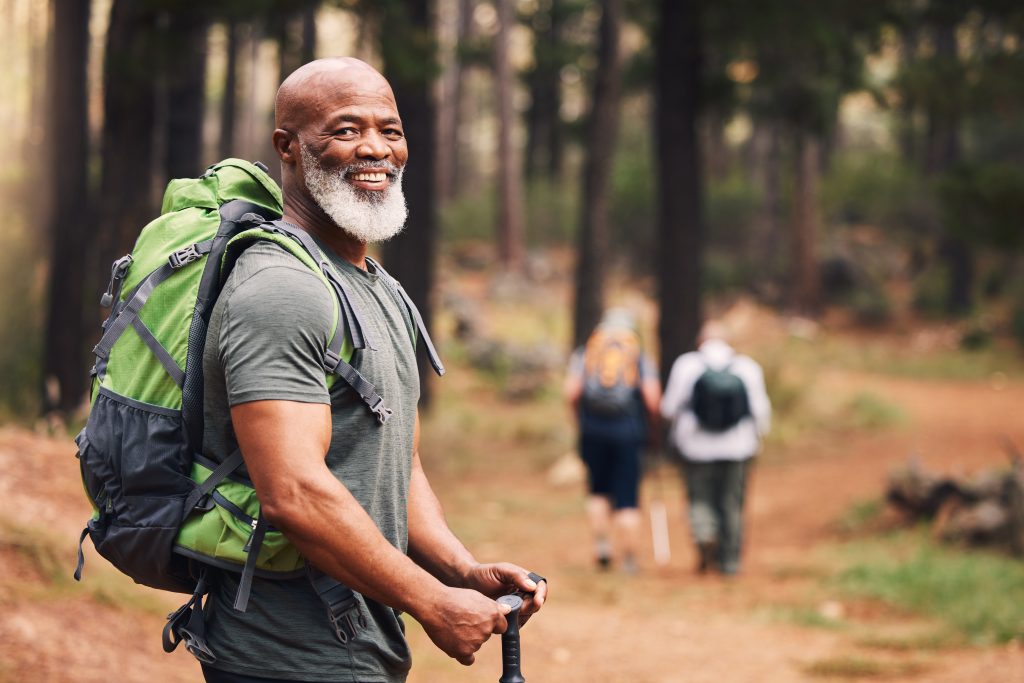 senior african american male wearing hiking backpack and holding hiking poles, smiling, with two unknown gender hikers in the background, out of focus